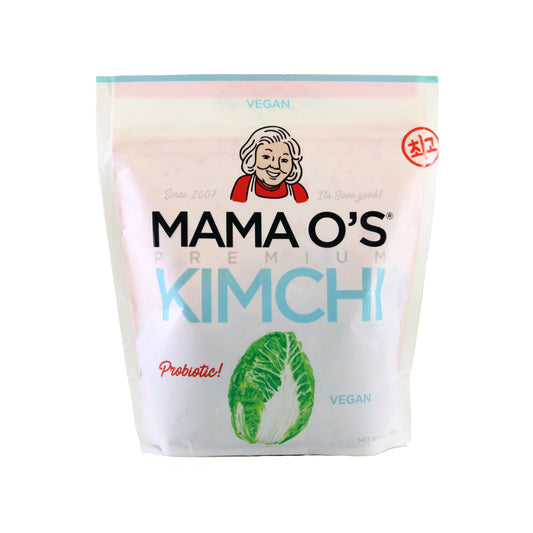A white pouch of Mama O’s Premium Vegan Kimchi, featuring a smiling woman and napa cabbage on the front with a “Probiotic!” label, showcases the probiotic benefits in every bite.