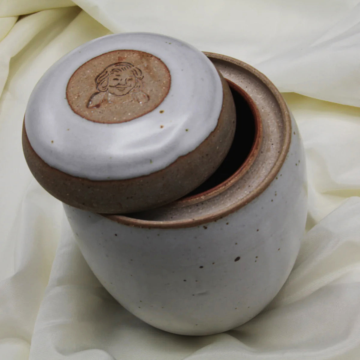 A custom, handmade Mama Os Homemade Kimchi Kit by Mama Os Premium Kimchi features a ceramic jar with a partial lid revealing a brown interior; the lid has an engraved smiling face, adding charm to any display.