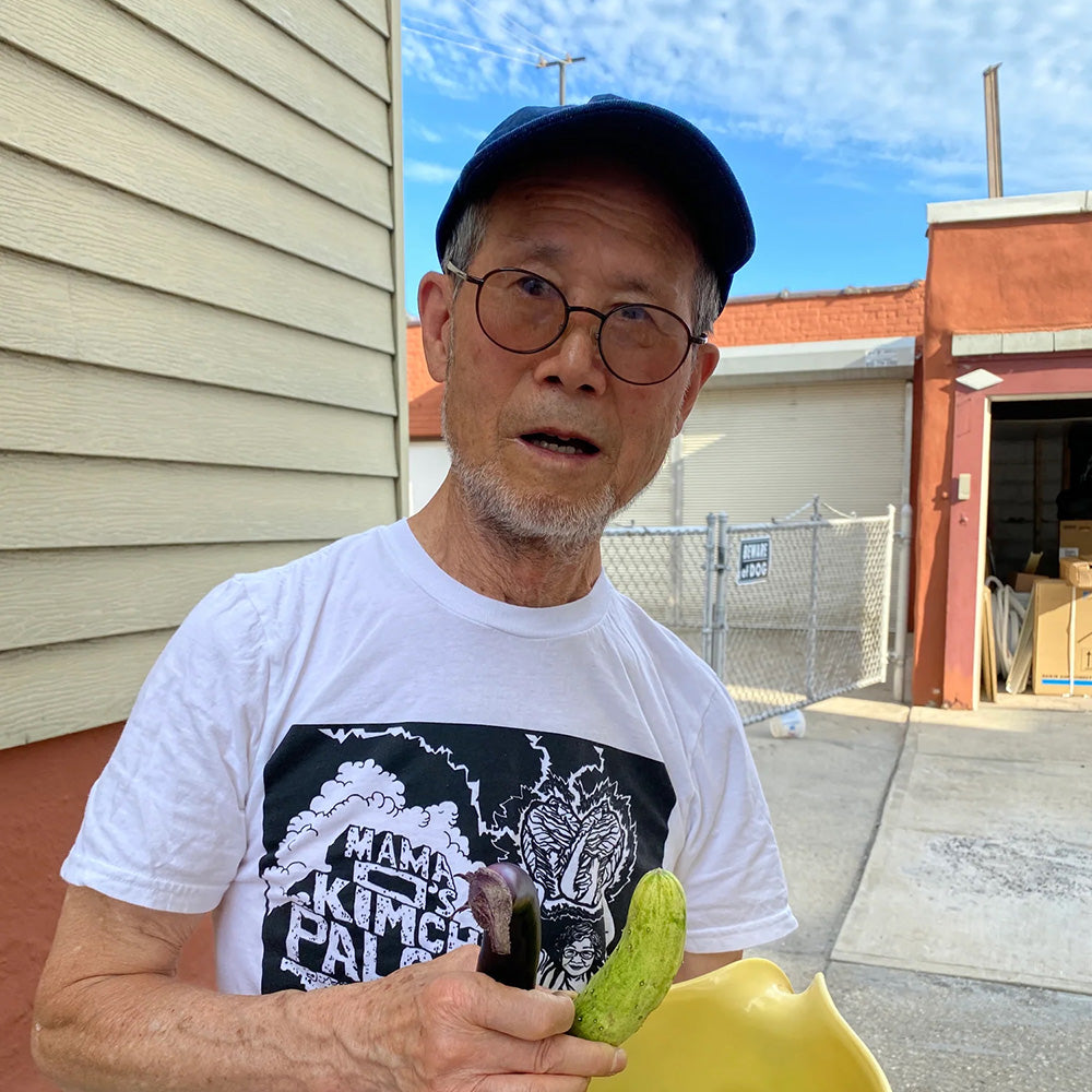 An older man in glasses, a blue cap, and a limited edition Mama Os Premium Kimchi Kimchipalooza X teeshirt stands outside holding a yellow bowl with cucumber and eggplant near a beige building under the blue sky.
