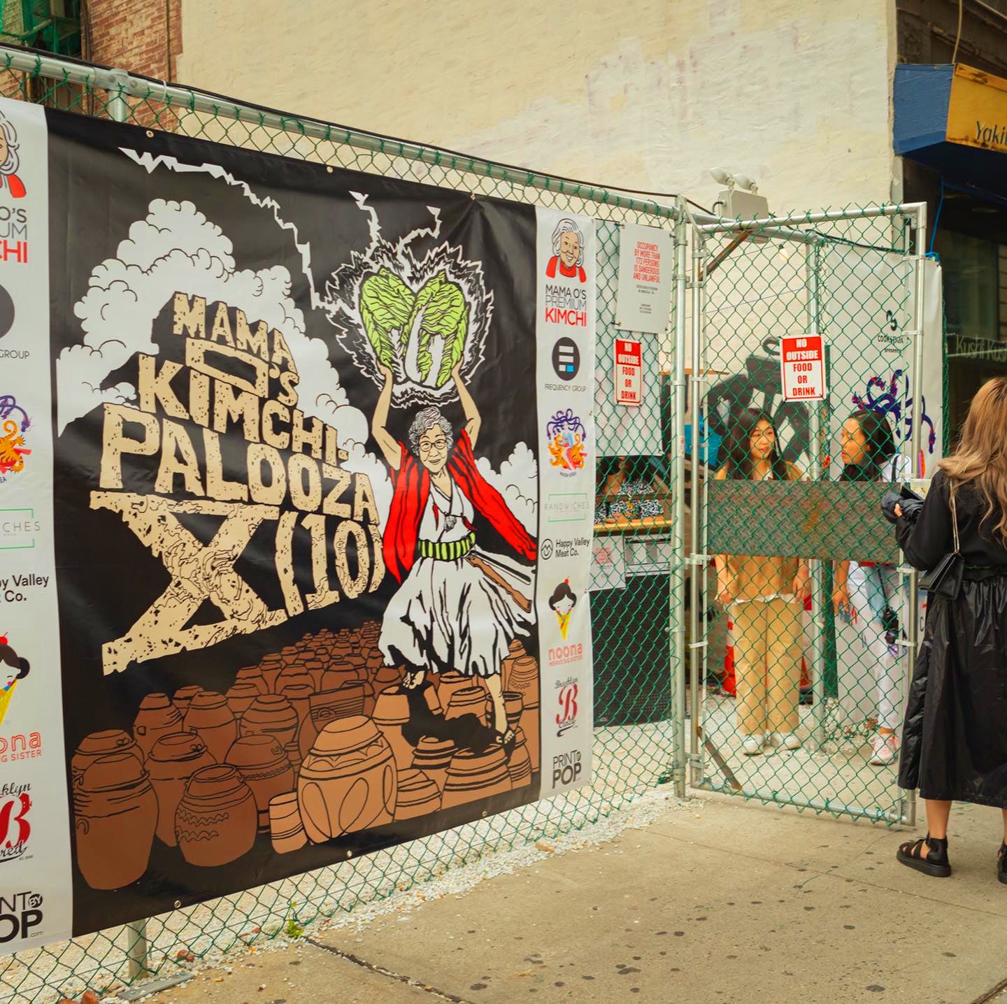 A large banner reading “Kimchi Palooza XII” hangs on a chain-link fence, featuring an illustrated person holding cabbages. People stand and walk nearby, with a brick wall and sidewalk in the background.