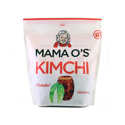 A white and pink pouch of Mama Os Original Premium Kimchi from Mama Os Premium Kimchi shows a cartoon woman’s face, cabbage and kimchi jar illustration, and text reading “Probiotic! ORIGINAL” on the front.