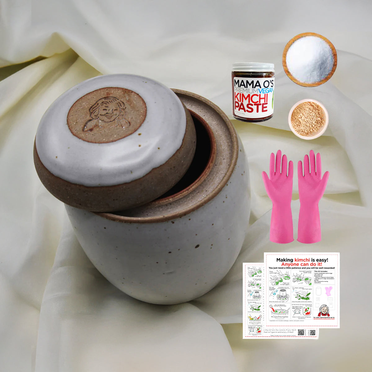 The Custom handmade Mama Os Homemade Kimchi Kit by Mama Os Premium Kimchi includes a ceramic hybrid onggi jar with lid, Magic Fermenting Powder, round filter, salt, pink gloves, and step-by-step kimchi instructions on white fabric.