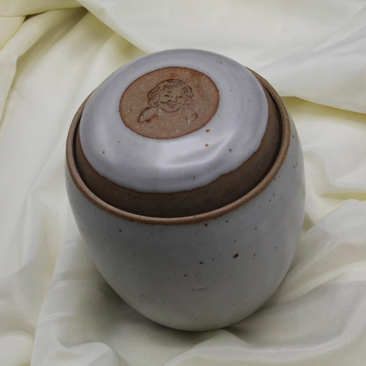 A round, speckled ceramic jar with a matching lid—engraved with a smiling face—rests on cream fabric. This charming vessel is part of the Custom Handmade Mama Os Homemade Kimchi Kit by Mama Os Premium Kimchi.
