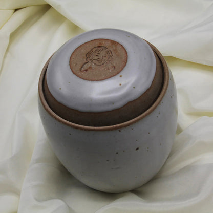 A round, speckled ceramic jar with a matching lid—engraved with a smiling face—rests on cream fabric. This charming vessel is part of the Custom Handmade Mama Os Homemade Kimchi Kit by Mama Os Premium Kimchi.