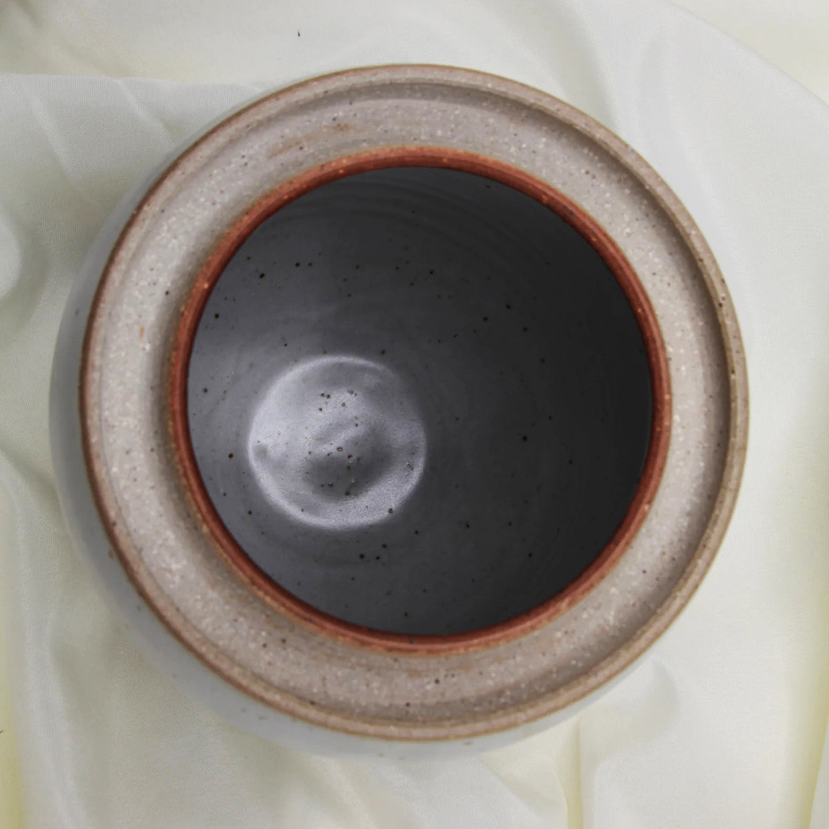 Top view of a custom handmade Mama O’s Homemade Kimchi Kit bowl by Mama O’s Premium Kimchi, featuring a dark matte interior and speckled beige rim, placed on light fabric.