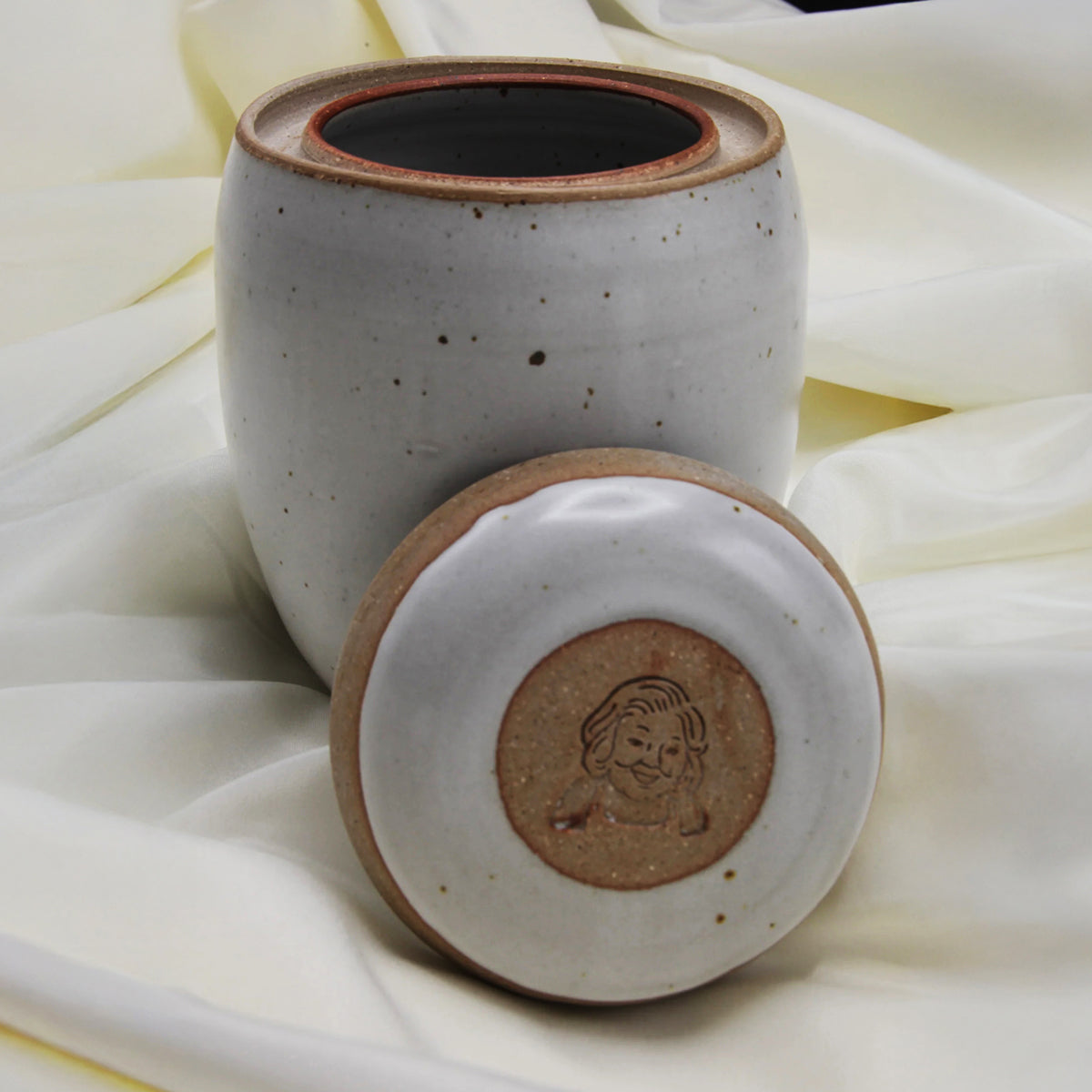 A Custom handmade Mama Os Homemade Kimchi Kit jar by Mama Os Premium Kimchi rests on cream fabric. Its lid, set in front, displays an engraved dog’s face. The jar’s white speckled ceramic glaze adds a stylish touch.