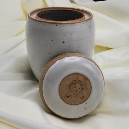 A Custom handmade Mama Os Homemade Kimchi Kit jar by Mama Os Premium Kimchi rests on cream fabric. Its lid, set in front, displays an engraved dog’s face. The jar’s white speckled ceramic glaze adds a stylish touch.